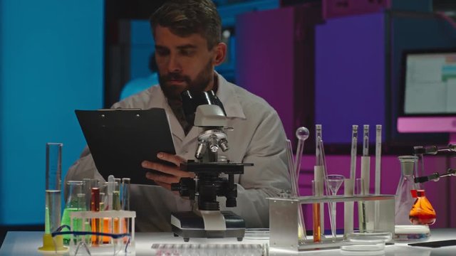 PAN of bearded male scientist checking data on clipboard and tablet, then looking at sample in microscope as his colleague working in background 