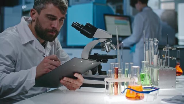 Bearded male scientist making notes on clipboard, then looking into microscope in laboratory 