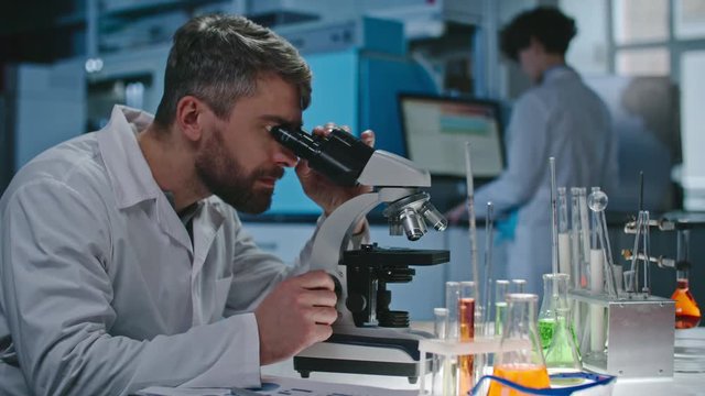 Serious bearded male scientist looking into microscope in laboratory, then making notes on clipboard 