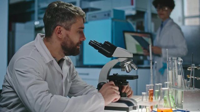 Male bearded scientist looking into microscope, then taking clipboard from female colleague and making notes