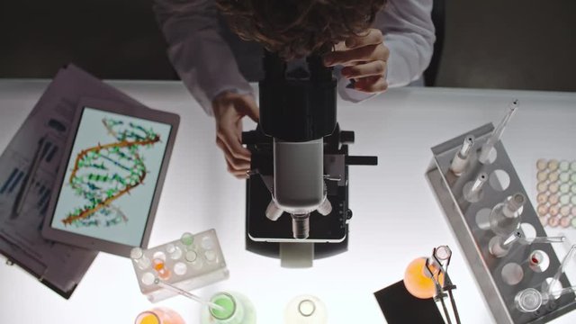 Top view of female scientist adjusting her microscope and looking at samples in laboratory, tablet with image of DNA lying beside