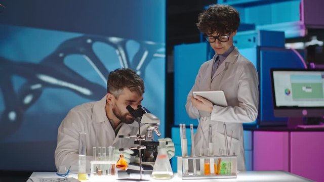 Male scientist looking into microscope in laboratory and discussing sample with female colleague standing beside him, projection of DNA on wall behind them