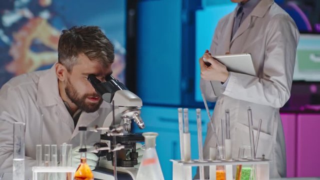 Tilt up of male scientist looking at sample in microscope and discussing it with cheerful female colleague standing beside him