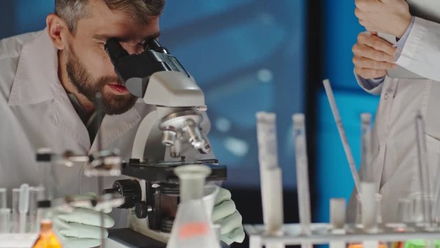 Tilt up of scientist with beard looking into microscope and discussing sample with female colleague making notes on tablet