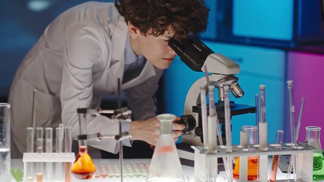 Curly female scientist adjusting lenses and looking into microscope in laboratory