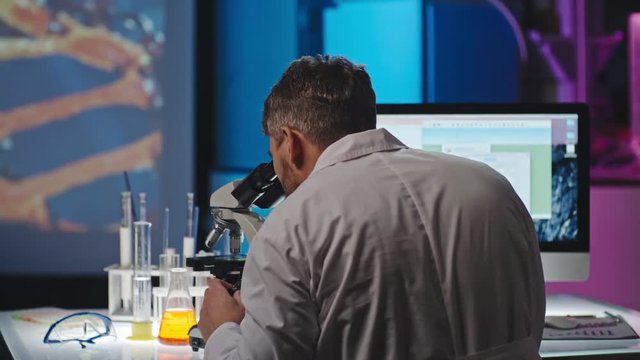 PAN with rear view of male scientist sitting in laboratory looking into microscope and making notes on computer, projection of DNA on wall in background