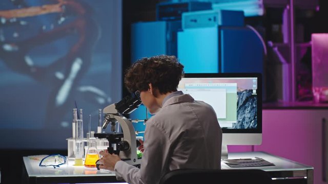 Wide shot with rear view of curly female researcher looking into microscope in modern laboratory, them making notes of computer