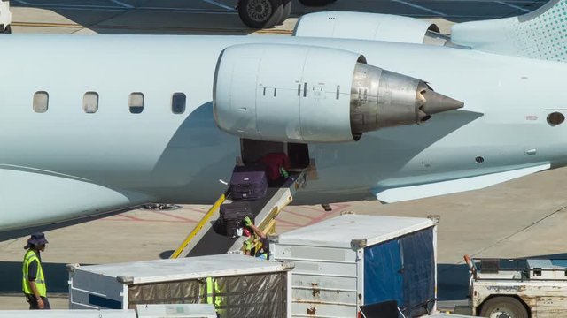Airport Baggage Handlers Workers Taking and Sorting Luggage Pieces from a Conveyor Belt Off of an Unknown Airliner and Loading it Onto a Cart on a Sunny Day