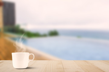 Hot coffee cup with steam on wooden table on blurred pool and sea background