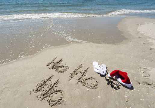 Ho Ho Ho written sand beach santa hat holly white thongs water edge.