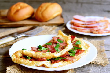 Ruddy bacon omelet. Fried omelet stuffed with bacon and parsley on a plate. Bacon slices on a plate, knife, fork, buns on old wooden table. Egg breakfast idea. Closeup