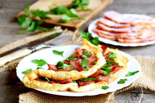 Delicious bacon omelette. Fried omelette with bacon and parsley for breakfast. Bacon slices on a plate, knife, fork, fresh parsley sprigs on old wooden table. Vintage style. Closeup