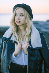 Obraz premium beautiful young blond woman standing by the sea at sunset wearing a black hat, a sheepskin coat and a white shirt. She is looking into the distance