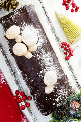 Buche de Noel on white marble with symbols of the season - a red linen napkin, holly berries, Christmas tree candle, and greenery with a small gift at bottom right.