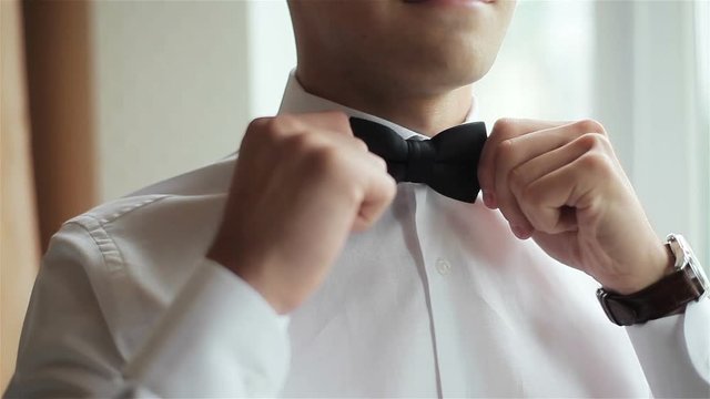 Dressing man straightens bow-tie close up. Well-dressed young man adjusts classic black bowtie on white shirt no face smile only. Male hands gets ready prepared for wedding correcting tie and image