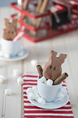 A hot chocolate cup with chocolate reindeer shaped and mini white marshmallows. A red stripy napkin underneath the cup and the sauce. A red metal caddy in the background. 