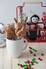 A big white ceramic cup filled with reindeer shaped marshmallow treat. A red metal basket with decor and treats in the background. Colorful candies scattered on the table 