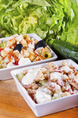 Two simple, fresh salads in white bowls on a wooden board next to cucumber and lettuce.