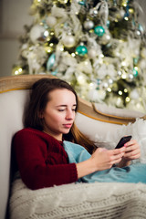 Young woman checking out her text messages on mobile phone as she liyng sofa in front of the Christmas tree at home.