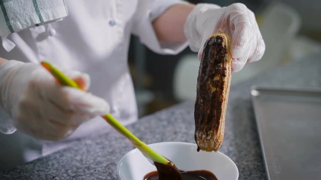 Cook pastry, in a white suit and gloves on his hands, smearing the melted chocolate yellow green silicone spatula on a freshly baked eclairs. Chocolate flows in a white bowl, which stands under eclair
