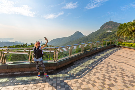 Young, smiling and fashionable tourist takes selfie on the popular free viewing terrace overlooking Victoria Peak Galleria that on the Reservoir in Hong Kong island.
