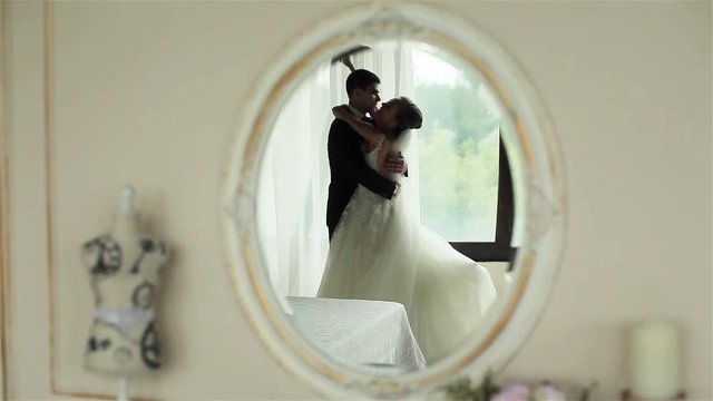 Happy newly wedded couple bride and groom embrace and kiss feeling happy reflected in mirror on wall of first night room interior. Happiness wedding honey moon marriage memories concept