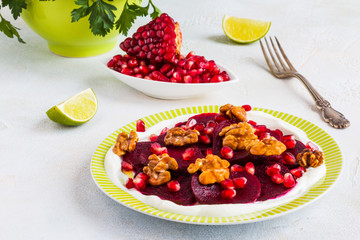 Dietary vegetarian salad of roasted beets with pomegranate seeds, walnuts caramelized in honey and natural yoghurt. Slices of lime, vintage fork, white and gray table, fresh parsley.
