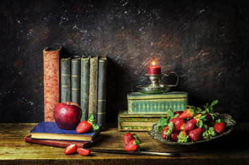 Classic still life with fresh strawberries,apple,illuminated candle and old books on rustic wooden background..
