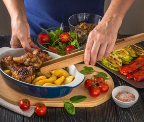 The cook prepares delicious and nutritious lunch with baked chicken drumsticks and potato in ceramic dish,  grilled vegetables, salad and tomatoes on the background of dark table