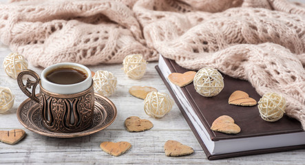 Cup of coffee and notebook with beige knitted scarf  and  wooden hearts lying on a white table