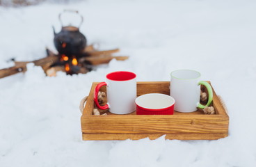 Romantic winter picnic. Two cups and a bowl on a wooden tray in snow. Copper kettle over an open fire on background, blurred. Boiling kettle on firewood. Lifestyle, camping.