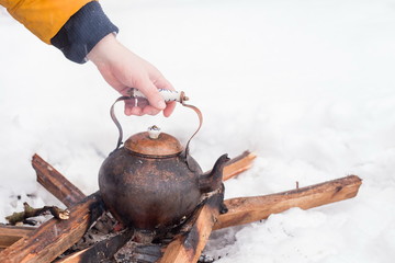 Hand of a man holding copper kettle over an open fire in winter. Boiling kettle on firewood. Open fire cooking. Snow around. Copy space. Lifestyle, camping.