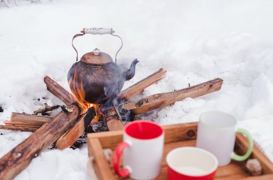 Romantic winter picnic. Two cups and a bowl on a wooden tray in snow. Copper kettle over an open fire on background, blurred. Boiling kettle on firewood. Lifestyle, camping.