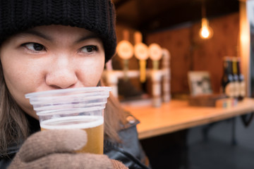 happy asian woman with black knitting wool hat holding cold craft beer in plastic cup, blur beer...