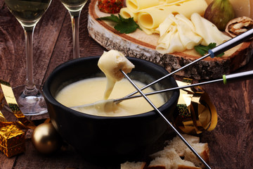 Gourmet Swiss fondue dinner on a winter evening with assorted cheeses on a board alongside a heated pot of cheese fondue with two forks dipping bread and white wine behind in a tavern or restaurant