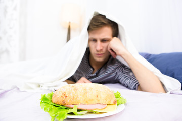 Sad and frustrated man lying and going to eat hamburger represented on plate. Single man lying on bed covered and looking at his breakfast.