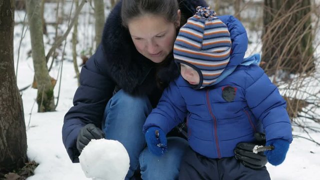 Attractive mother and baby sculpts small snowman in the winter forest. Family fun for the Christmas holidays