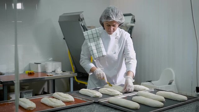 The woman in a candy store. The brunette in a white form does preparations for roasting of bread. The girl of middle age has created a form of bread from the test.