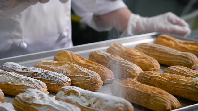 Pastry chef, dressed in a white chef's suit and transparent gloves sprinkle with powdered sugar from a special cup - a sieve to cook eclairs, standing on a metal tray