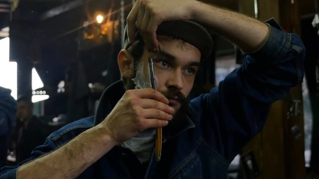 Handsome man shaving in barbershop with foam and manual dangerous razer. Young barber shaves himself