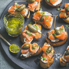 Homemade salmon crostini with cream-cheese, watercress, capers and pesto suace in round black slate stone plate over grey background, selective focus, square crop