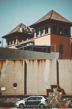 Vertical photo of prison in Barcelona with two pyramidal towers, big concrete rusty wall, police car, no stopping  road sign, sunny summer day, Spain