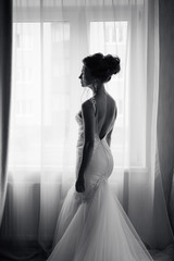 Silhouette of a beautiful bride in a traditional white wedding dress, stood by window.