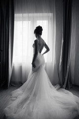 Silhouette of a beautiful bride in a traditional white wedding dress, stood by window.