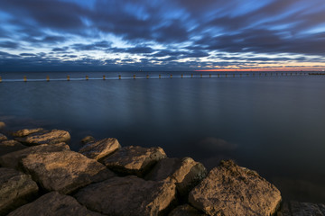 A shot of an empty lake taken from a pier during the sunrise with Chicago's Skyline visible in the background. Beautiful rocks are present in the foreground.
