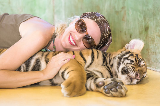 Smiling beautiful woman with sunglasses, embraces a little tiger, Panthera Tigris, lying in Thailand. Concepts of courage, fun and dangerous.