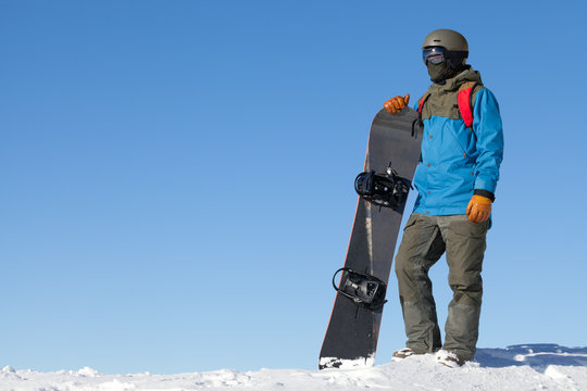 Male snowboarder taking a look at landscape at the top of mountains with blue sky on background