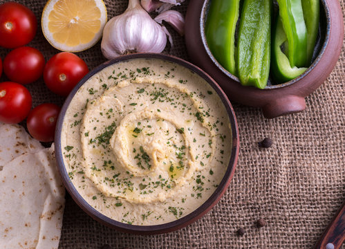 Humus in homemade bowl with vegetables on jute background. Top view.