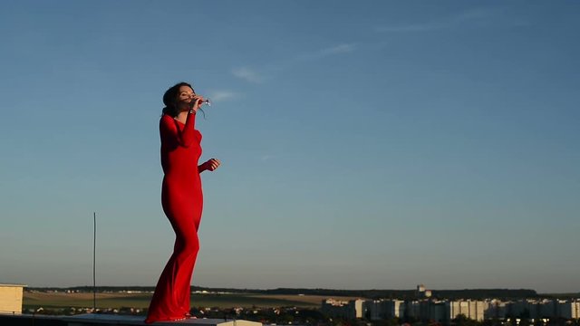 young beautiful brunette woman in a red dress with a glass in his hand against the blue sky on the roof