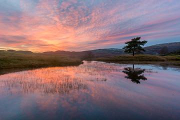 Fototapeta na wymiar Dramatic fiery orange sunset with mirrored reflections in water at Kelly Hall in the Lake District.